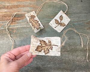 This shows three birch bark accent tags with fall leaf artwork burned onto the bark. Each tag has a piece of twine tied to it. 